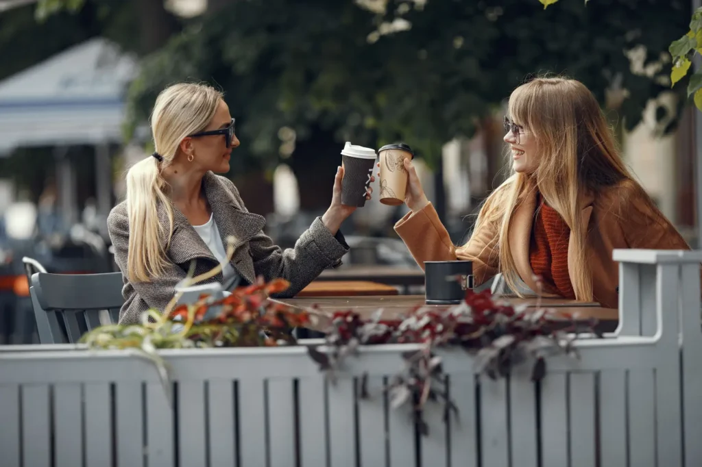 Two smiling women enjoying coffee together while sitting outdoors at a café.